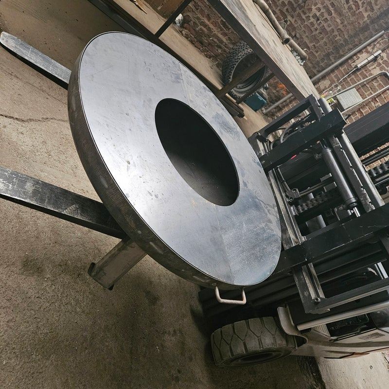 Grand anneau plancha en acier pour brasero, soulevé sur les fourches d’un chariot élévateur à l’atelier
