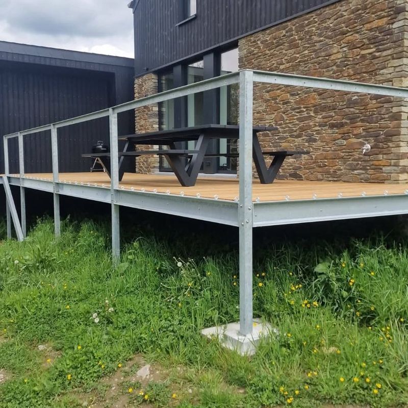 Terrasse sur pilotis en acier galvanisé avec garde-corps, plancher en bois, devant une maison à bardage noir et pierre