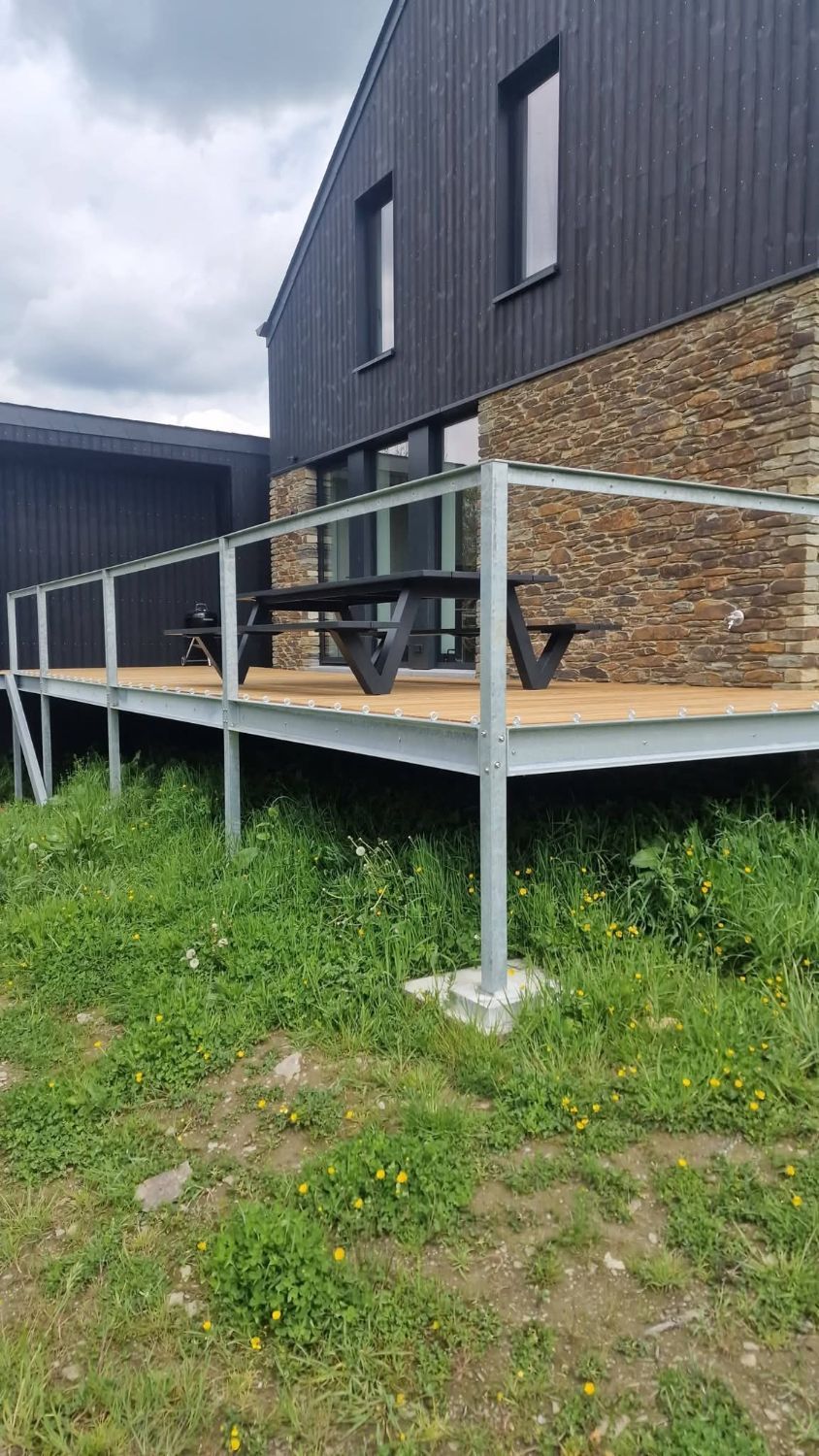 Terrasse sur pilotis en acier galvanisé avec garde-corps, plancher en bois, devant une maison à bardage noir et pierre